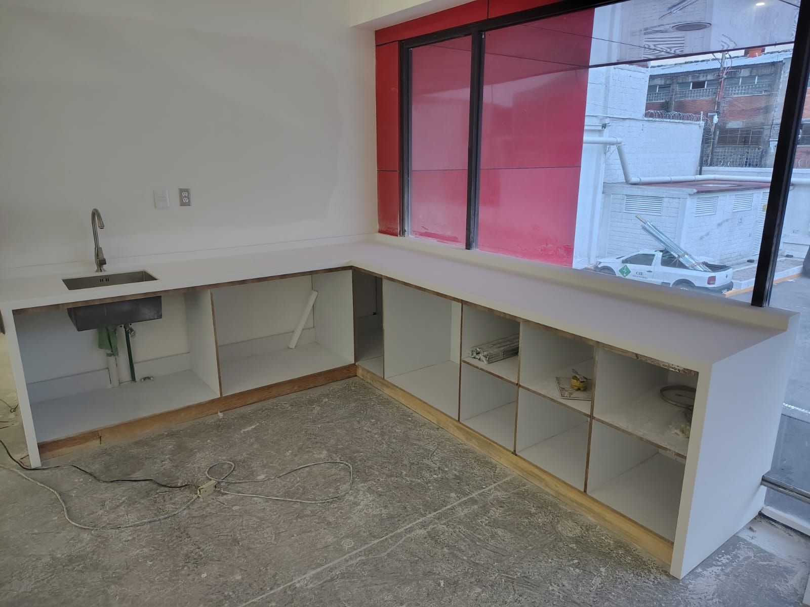 Cubierta blanca en L con tarja de acero sobre muebles abiertos junto a un ventanal